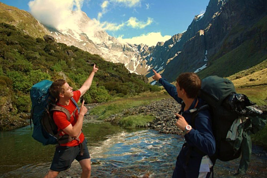 Two enthusiastic hikers strike a pose at a valley river crossing. They are turned away from the camera and pointing up towards a snow capped peak.