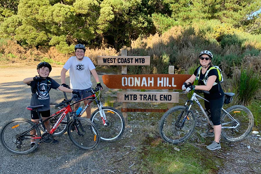 A woman with her two sons, all on mountain bikes, by some signage at the end of Oonah Hill trail. They are all smiling at the camera.