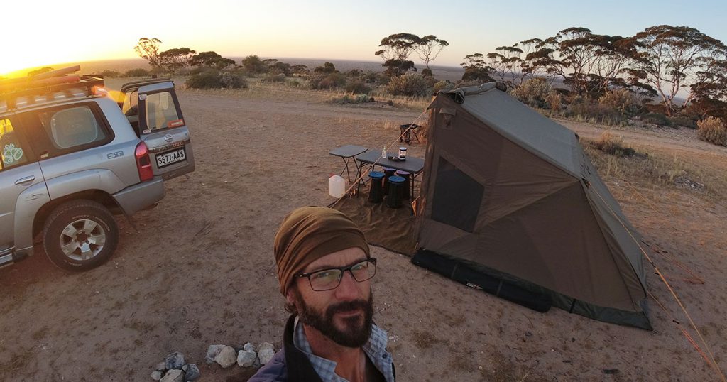 Ben is standing in front of his 4WD and neat tent set-up at sunset. He is wearing a brown headscarf.