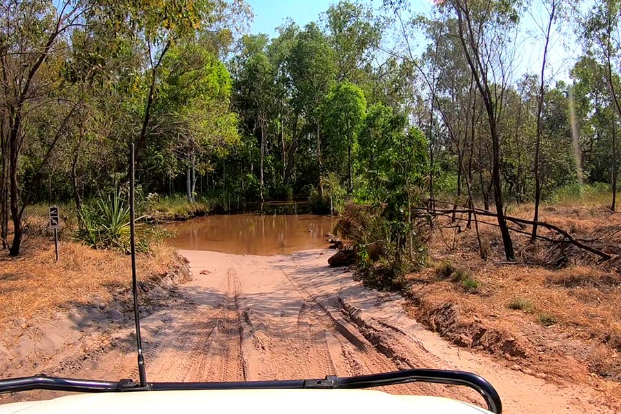 A sandy track in the Top End of Australia with a 4WD vehicle approaching a wide creek crossing. Image is taken from the driver's perspective and there are trees and foliage all around.