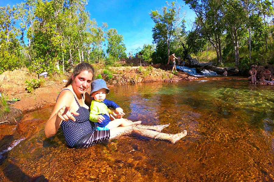 A woman in bathers sits in a rockhole, cooling off with a young toddler in her lap. She's half smiling and pointing at the camera with a few scattered people in the background also sitting in the water. The river bank is behind her with trees and blue sky.
