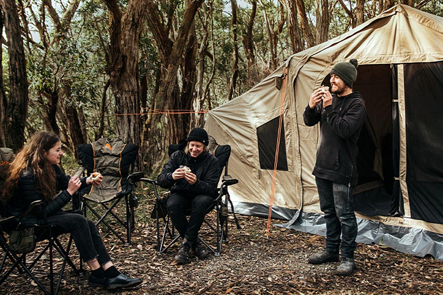 Outside their tent, two campers are sitting in camp chairs and one is standing nearby. All three are eating lunch and chatting. The campsite is green and leafy, and the campers are rugged up in dark clothing.