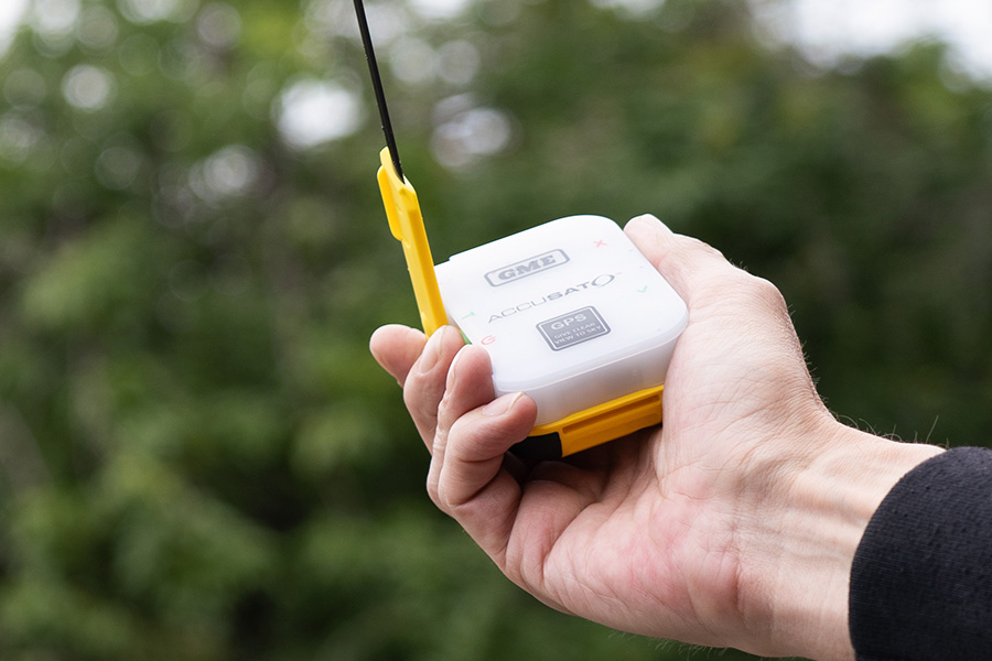 A hand is holding a GME Accusat PLB with yellow detailing. The backdrop is leafy and green.