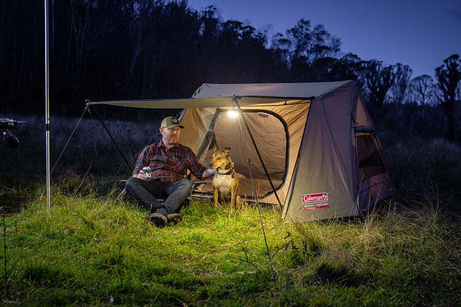 It is evening time. The Coleman Instant Swagger Tent is erected on a bed of soft, green grass with the awning extended. A man dressed in a red flannelette shirt is sitting in lamplight with his dog by his side. In the backdrop is a forest of trees. 