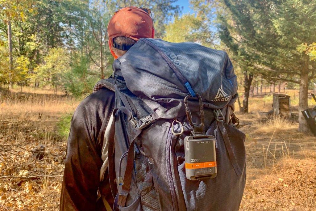 A man with a red cap is standing with his backpack to the camera in a hot, dry environment, with a Spot satellite communicator hanging off a carabiner attached to his pack.