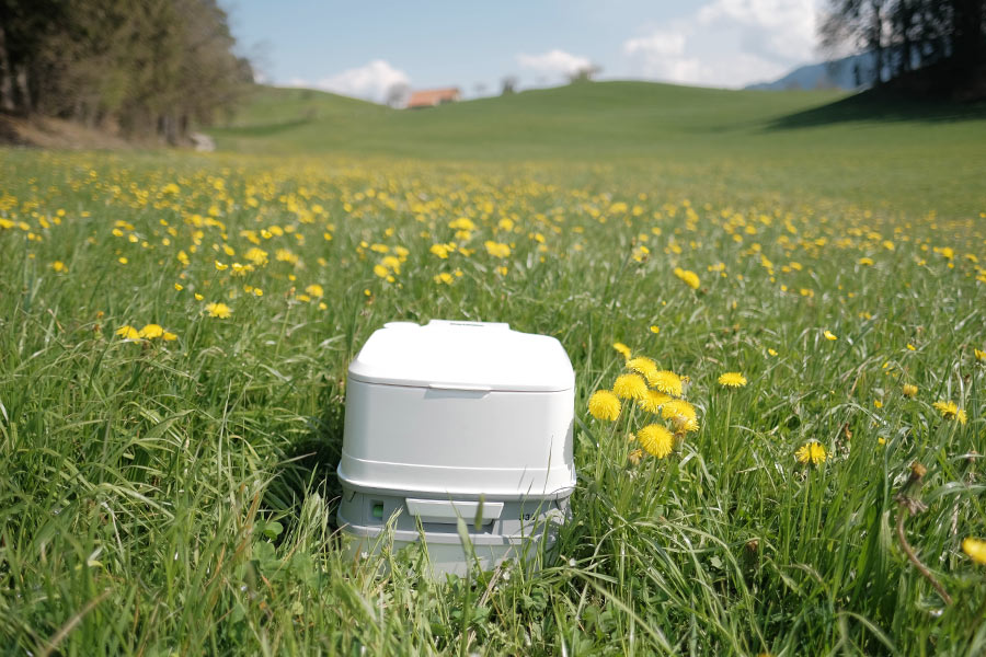 A white, stout, portable toilet sits in the middle of a green field dotted with yellow dandelions.