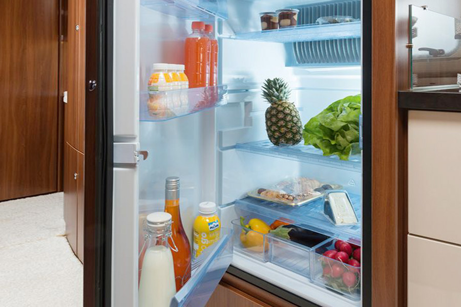 A caravan fridge door is ajar, presenting fresh produce and condiments stacked on its shelves and in its drawer compartments. The space is well lit with bright, natural lighting.