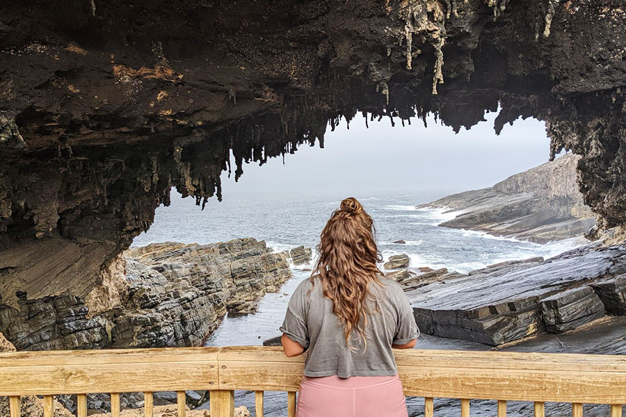 Lauren stands at Admirals Arch in pink sports shorts and a grey t-shirt, her brunette hair out and flowing. She surveys the grey, misty morning and the ocean through a large rocky arrch.