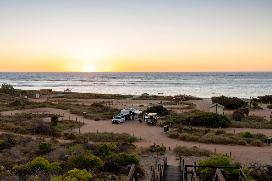 Two groups of 4WDs and camper vehicles are parked in a beach carpark, overlooking the shorelines. The sky is a dusty orange-pink topped with cool blue, reflected in the ocean. There are flat, sandy pathways snaking throughout, and patches of green shrubbery clustered off the tracks. It seems to be either sunset or sunrise. 