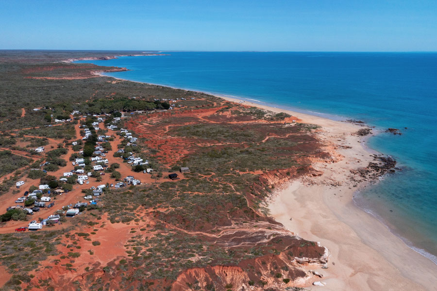 A birds-eye-view of Barn Hill Station shows rich, red land against vivid blue ocean, the white sand of the beach padded in between. On the land, there are three rows of cars, caravans, and camper trailer set-ups.  