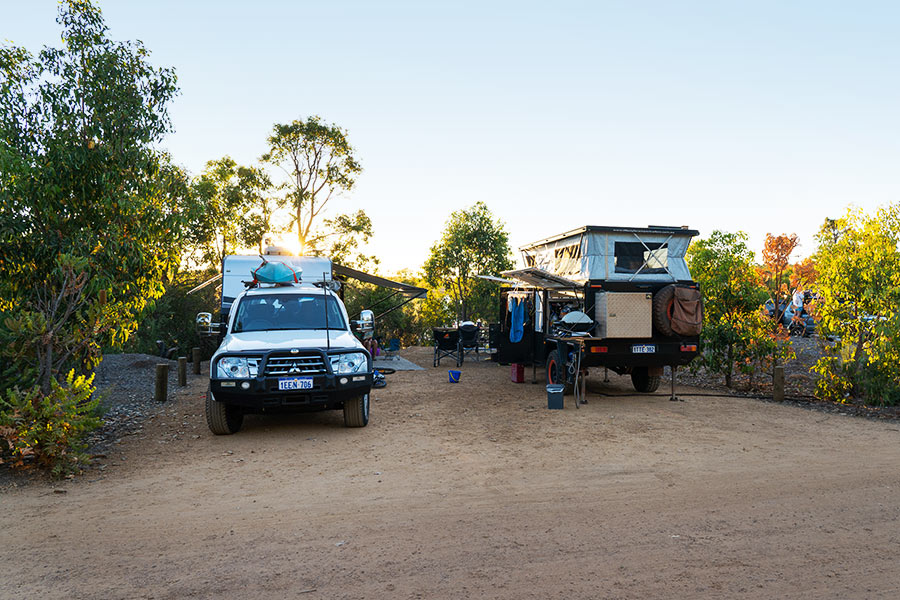 A 4WD and camper trailer are set up alongside each other in a campsite. The sun is peaking through the trees, and the sky is a light, dusty blue. The ground is flat with fine white gravel, and the trees nearby are a vivid yellow-green.