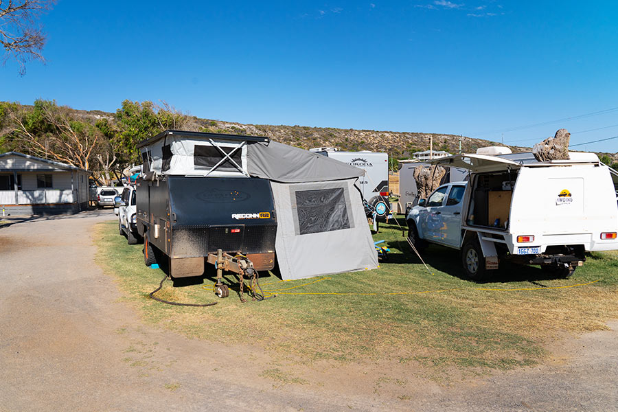 A 4WD and camper trailer are set up alongside each other in a busy campsite, bordered by a dusty driveway. The sky is a vivid blue, suggestive of the mid-morning or mid-afternoon. The ground is flat with maintained, yellow-green lawn, and in the background are trees and shrubbery. 