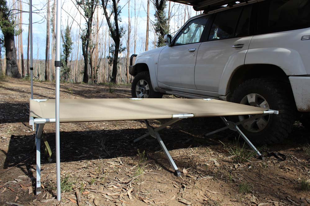 A camp stretcher sits among the soft dirt and gum leaves beside a white 4WD underneath the awning. In the background are tall, leafless trees, and a blue sky padded with clouds. The stretcher is a pale, dusty shade of brown.