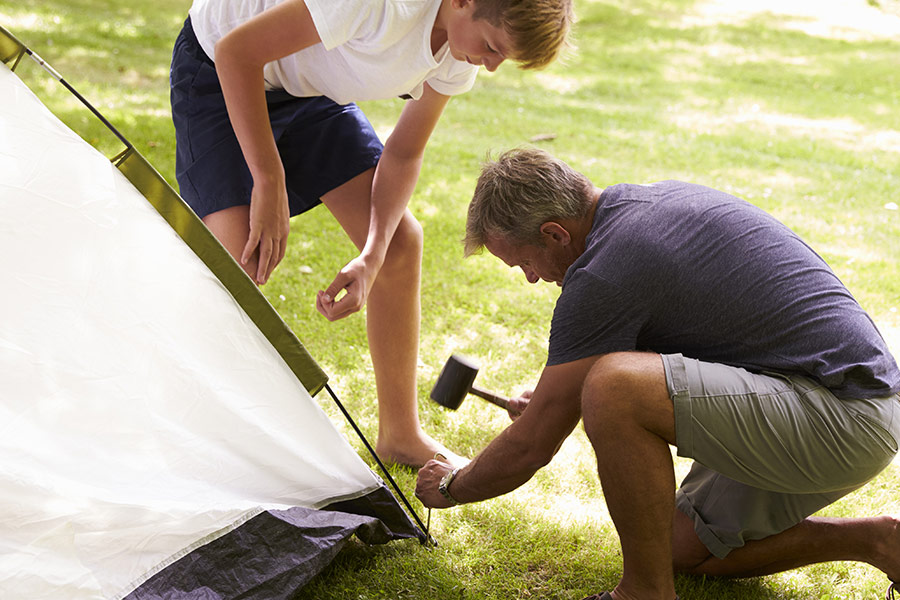 A man is crouched to the floor with his teenage son are tapping a tent peg with a mallet into the corner of their tent. The man is wearing a navy blue shift and grey shorts, and his son is wearing a shite shirt with navy shorts. The lawn is a bright, yellow-green. It is bright, broad daylight.