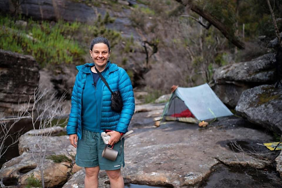 Kate stands on a slab of rock wearing a blue puffer jacket and teal-blue shorts, holding a mug. Behind her is her small hiking tent and some green shrubbery. She is camping by a creek. 