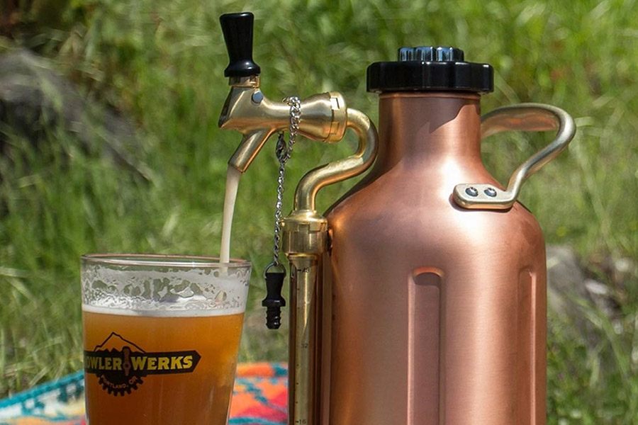 A bronze growler is positioned next to a glass of golden beer, pouring foamy brew from its spout. The top of the glass is foamy, with a logo printed on the outside facing the camera. It is daytime, and the backdrop is vivid green shrub. 
