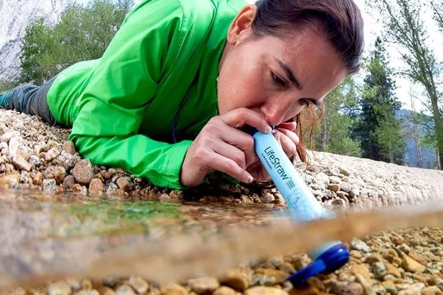 A lady in a green, long-sleeved shirt is lying flat on her stomach at the pebbly edge of a creek, drinking water through a straw filter. It is daytime. The water is clear.