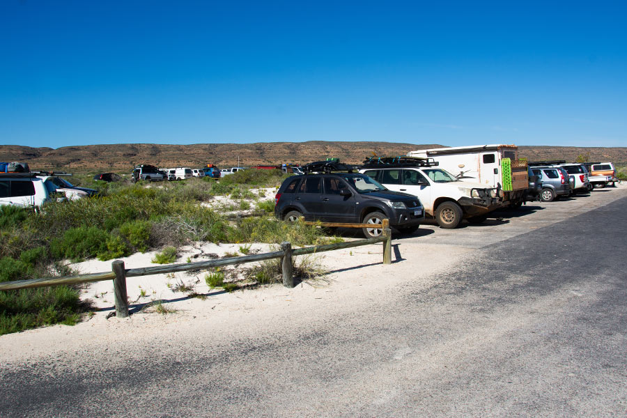 A beachside carpark is packed with cars filling every space. The bitumen is dusted with white sand, the sky is a vivid blue, and green shrubbery is clustered amongst the sand. 