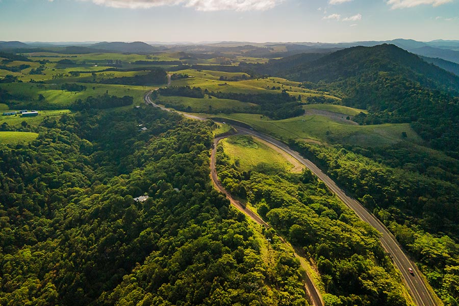 An overhead shot of the Atherton Tablelands shows shades of vivid yellow greens and darker patches, trees clustered together and roads scribbling their way through the landscape. The sunlit patches are golden and the mountains in the background are shades of blue.