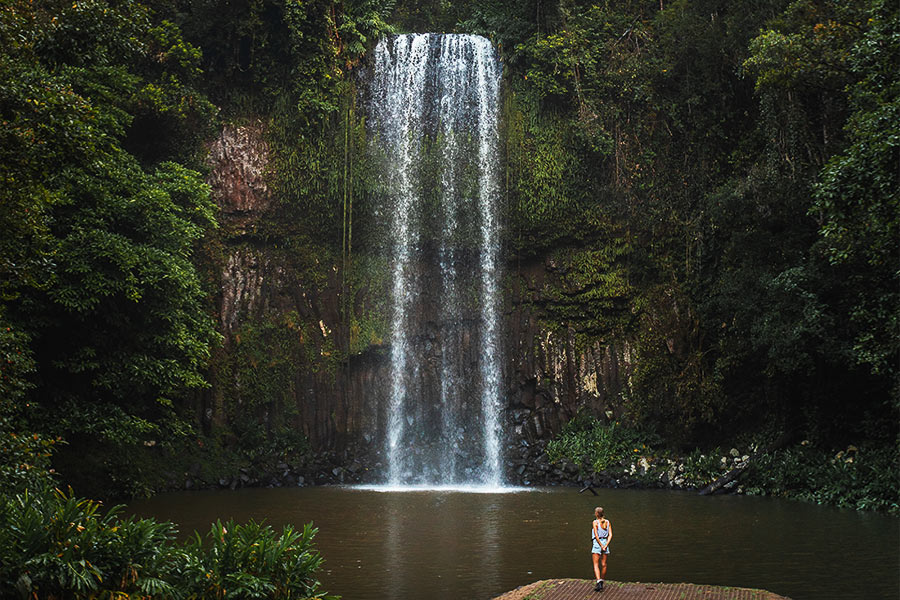 A girl is standing on a platform overlooking a body of water, looking up at a waterfall tumbling down against the rockface.