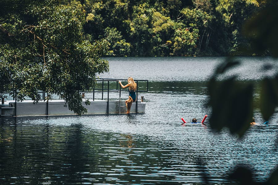 A girl in a blue swimsuit is climbing from the lake up the ladder of a jetty. There are overhanging trees, and the water is a deep blue-green.