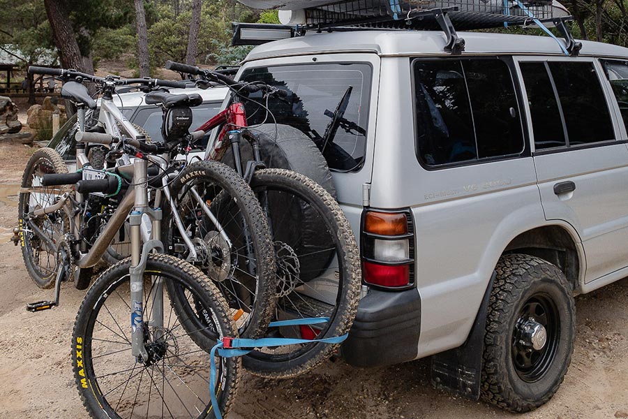 Three mountain bikes are stacked on the back of a four-wheeled drive, protruding outwards from the spare tire and secured by a blue strap.