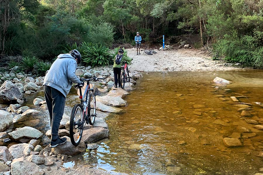 Three cyclists are wheeling their bikes over the rocks the create a stepping-stone bridge across a yellowing, shallow creek.