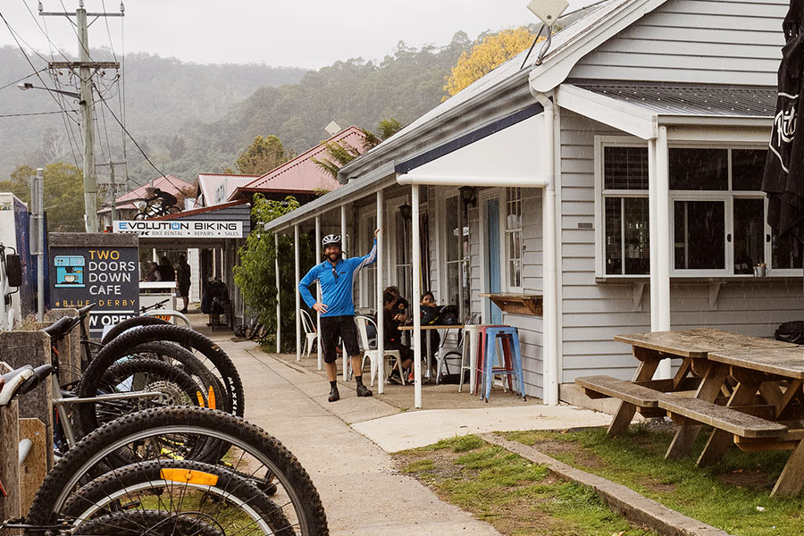 There are a range of MTB businesses in Derby for shuttles, guided tours, bike hire, and servicing. A man in a blue jacket is standing outside a small, picturesque cafe built with white slates of wood and blue, red, and white stools perched out the front. There is a bike rack holding multiple bikes with their wheels positioned to the sky.