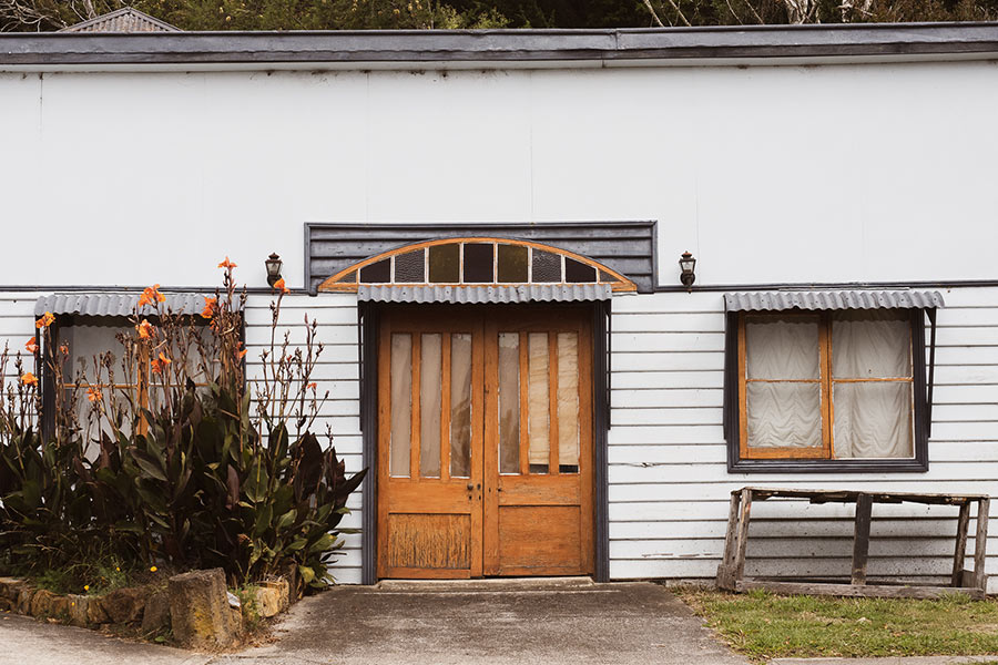 I took a walk with my camera to capture the history and character of this thriving little town. A picturesque white building stands with double wooden doors and white wooden slates lining the front wall. To the left are large flower arrangements with orange flowers on the end of long stems. The window roofs are corrugated iron.