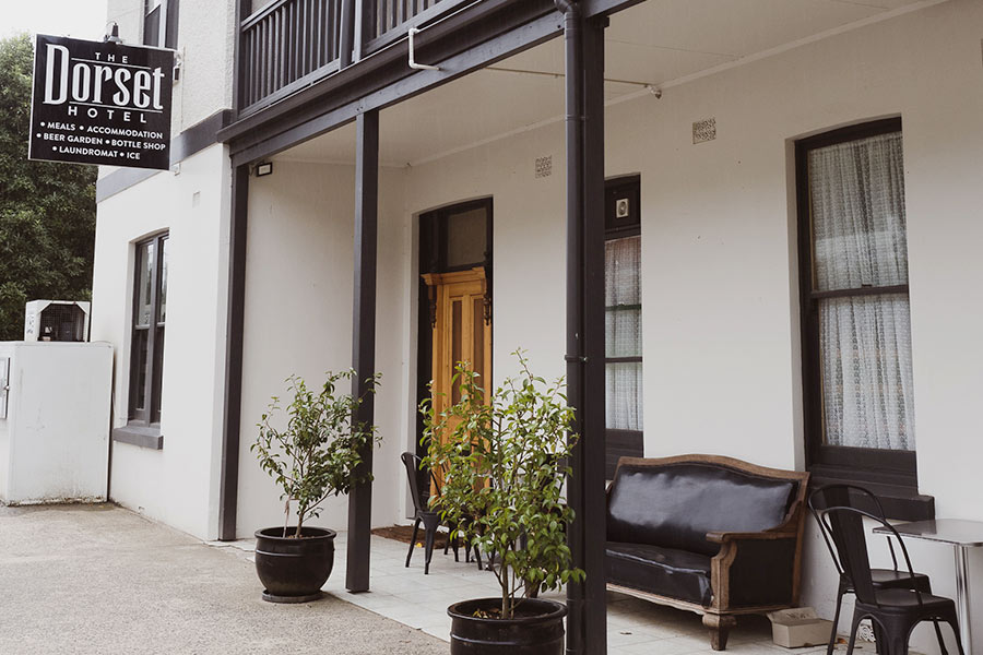 A white building called The Dorset Hotel - black trims along the balcony and doorways, with a black leather couch and black steel chairs outside. Pot plants are positioned by the supporting posts, growing lush green bush. 