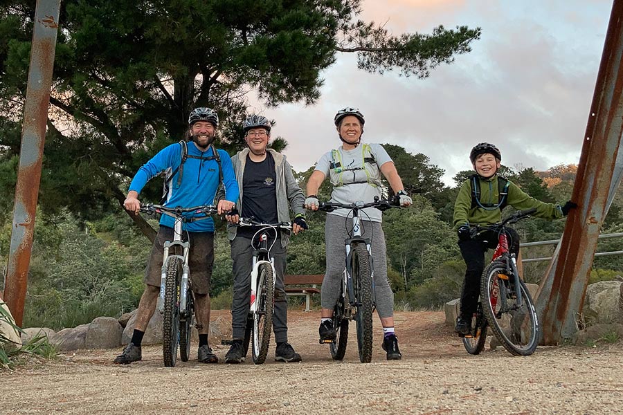 After the West Coast, we set our sights eastward to Derby for a very different experience. A family of four is perched on their bikes on a rubbly surface, with a pink stippled sky and trees behind them. The father is wearing a blue jacket, the second son wears a navy t-shirt under a grey jacket, and the mother wears a pale grey-blue t-shirt. Each family member is wearing a helmet.