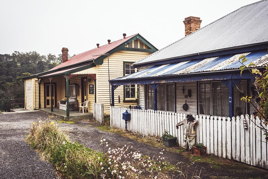 Derby was originally a tin mining town, having experienced boom, tragedy, and eventual decline. Two pretty houses of Derby sit side by side, the house on the right lined with a white picket fence. The house on the left is yellow with a red roof and green trims. The house on the right has dark blue trims, and the paint on the roof flaking away.