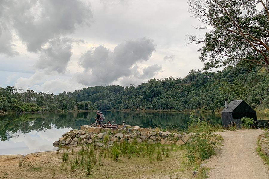 Two bike riders are parked on the rocky edge of a lake. The lake is glossy, reflecting the bushy, green forestry. There are vivid yellow-green grasses growing in patches on the side of the bike path. 