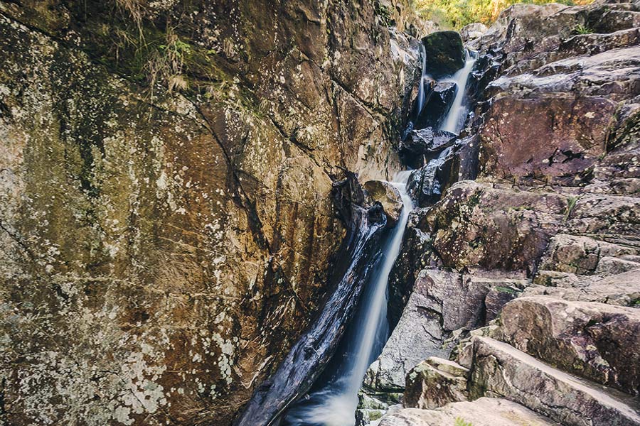 A rushing waterfall travels down a steep, sharp, faceted rockface. The rocks are marbled, coloured with browns, golds, moss-greens, and stippled with white. 