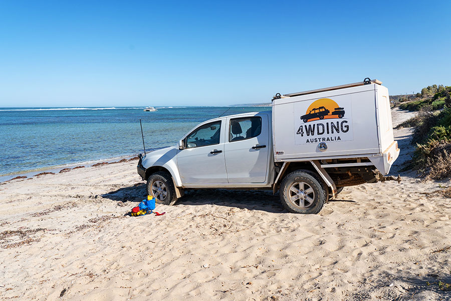 A 4WD with a logo reading '4WDing Australia' is parked on the soft, white sands of a beach. The sky is a two-colour gradient of blue, reflected in the ocean. It appears to be the middle of the day. 