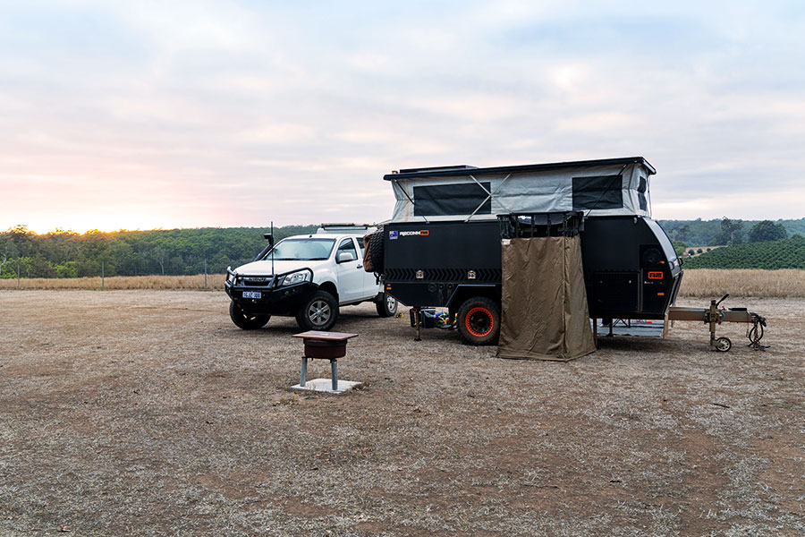 A 4WD and camper trailer are set up alongside each other in the countryside. The sun is melting over the horizon and the sky is a light, dusty blue, padded with clouds and tinged with pink from the sunset/sunrise. The ground is flat and dusty with stubbly, dry grass.