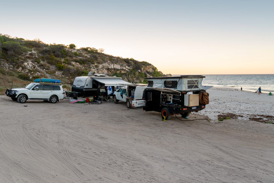 Two 4WDs each with a camper trailer are parked perpendicular to each other on the white sands of a northern Australian beach. The sky is a dusty pink topped with cool blue, reflected in the ocean. There are children small shapes suggestive of humans swimming in the water in the background. It seems to be either sunset or sunrise.