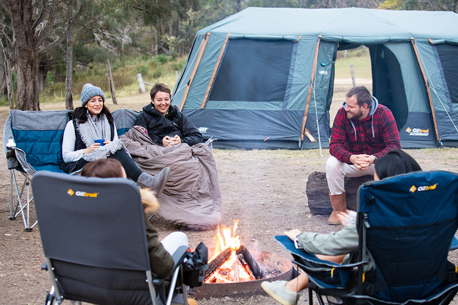 A group of campers are sitting around a campfire with a tent in the backdrop.