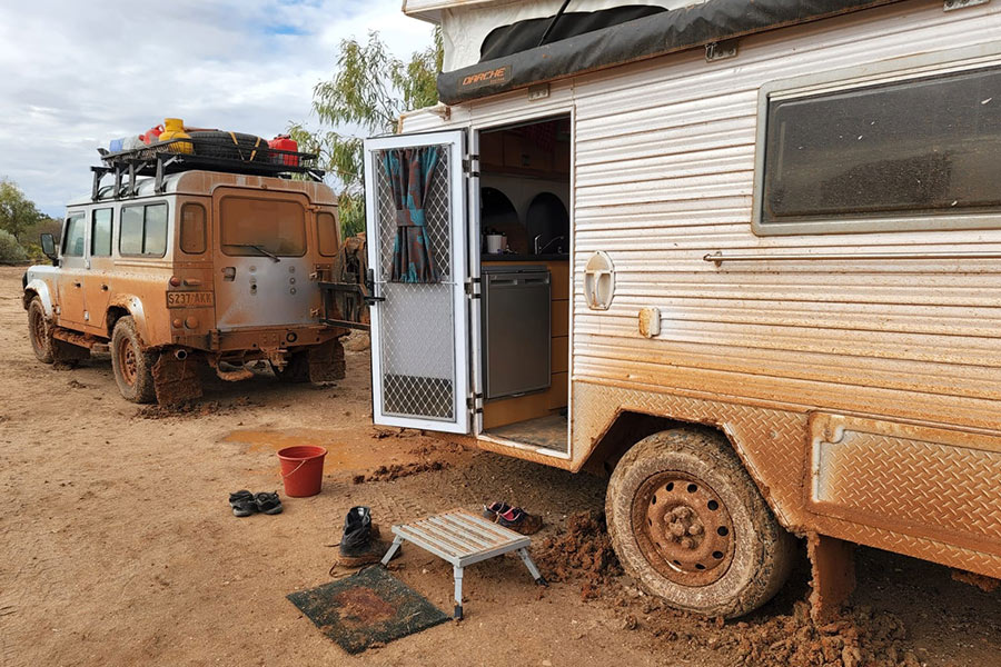 A caravan and 4WD are parked on red dirt, covered in red mud.