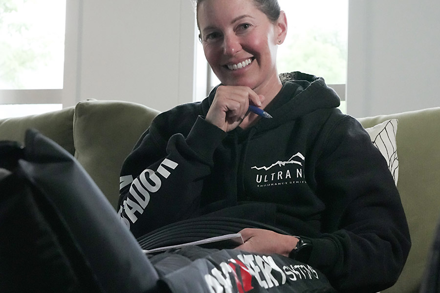 Tanya is sitting relaxed on her couch with her legs up and feet in her compression boots. She is wearing a black hoodie with white text, a big smile on her face, and holding a pen up by her chin. 