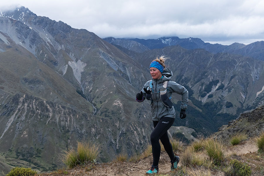 Tanya is running along a narrow trail against a vast, mountainous backdrop. The grooves of the ridges, ranges, and valleys are visible on the mountain face, and their tops are capped by a thick mist. Tanya wears black running tights, bright blue runners, a brightly patterned, blue headband, grey jacket, and hydration vest. 