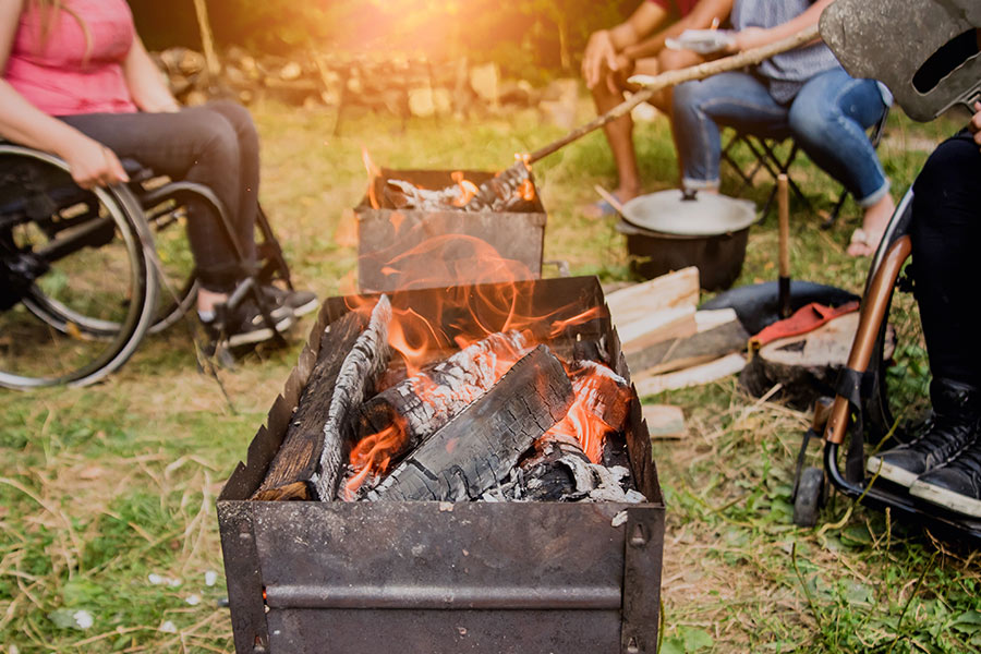 A fire pit filled with ashy wood, hot amber coals, and a bright orange flame sits in the corner. There are campers sitting nearby in the background, including an individual in a wheelchair wearing a pink shirt.