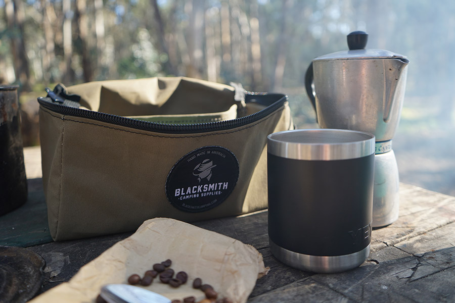 A Blacksmith Camping Supplies coffee kit bag sits on a wooden table,  unzipped. Beside it is a percolator, insulated mug, and some coffee beans resting on their brown paper bag.  