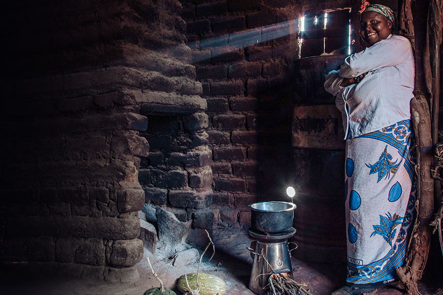 An dark-skinned woman is standing in a dark, brick room by a fireplace, wearing a long, blue, patterned skirt with her arms folded across her chest. She is smiling, and beside her is a cooking pot on top of a BioLite stove. 