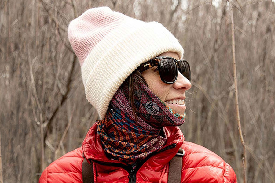 A woman wears a colourful, patterned neck scarf with sunglasses, a cream and pale pink beanie, and a red puffer jacket. She is looking to her right. 