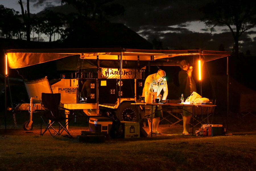 A vehicle camp setup is illuminated by a vivid orange glow. Under the awning is a man preparing food at a table, an Esky, and some deck chairs. 