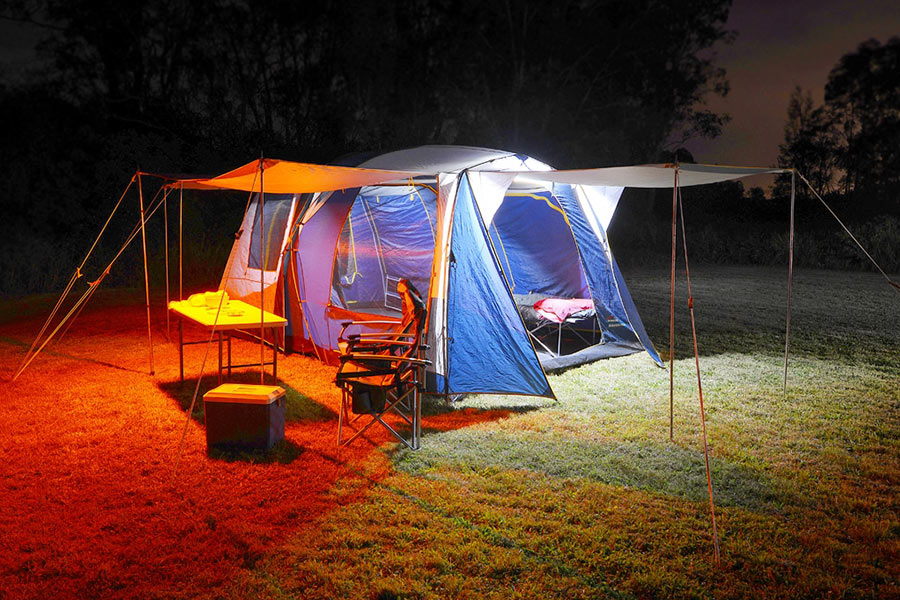 A dome tent is illuminated on two sides. One side emits a vivid orange glow, while the other glows a bright white. Under the awning is a table, an Esky, and some deck chairs. 