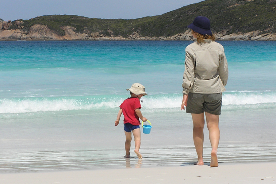 A woman and a child are walking towards the ocean on a sandy beach. The child carries a blue bucket. 