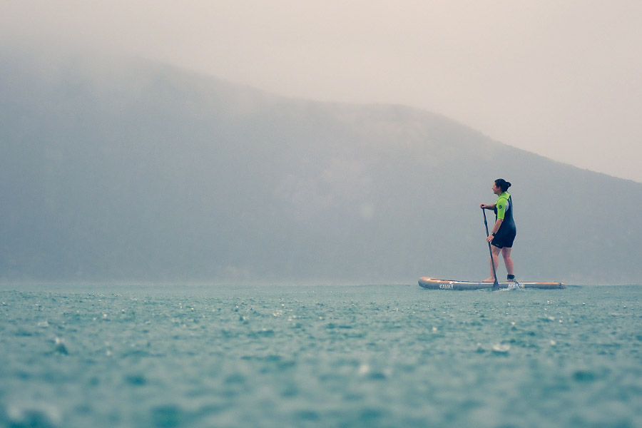 A woman is SUP-ing in the rain, on the ocean. 
