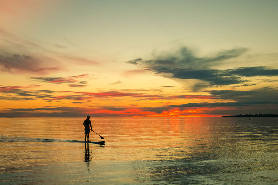 A man is SUP-ing on the ocean at sunset. The sky is a vivid red and golden light melts on to the water's glassy surface. 